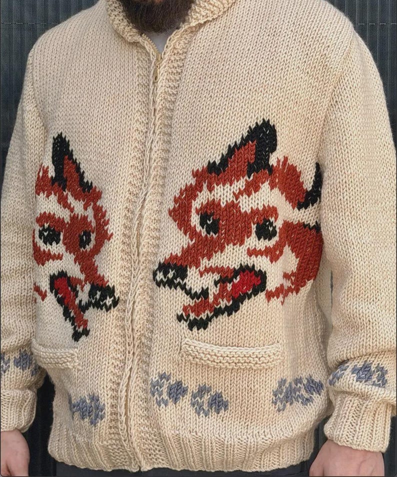 May include: A beige knit zip-up sweater featuring two large fox motifs on the front panels. The foxes are rendered in orange, black, and white yarn. The sweater includes two front pockets and a row of small grey paw print patterns along the hem.