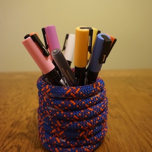 May include: A blue and orange rope pencil holder with a variety of coloured markers inside. The markers are pink, purple, white, yellow, blue, and black.