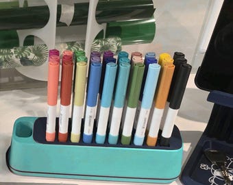 May include: A teal pen holder with a navy blue interior holds a collection of colorful markers. The markers are in various colors, including pink, orange, yellow, blue, green, and black. The background includes rolls of green and white patterned vinyl.
