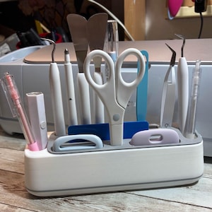 Può includere: Un organizer bianco e grigio per attrezzi artigianali pieno di vari strumenti, tra cui forbici, pinzette e spatole. L'organizer ha una forma rettangolare con un bordo grigio e diversi scomparti.