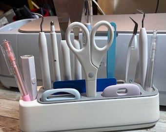 May include: A white and gray craft tool organizer filled with various tools, including scissors, tweezers, and spatulas. The organizer has a rectangular shape with a gray trim and several compartments to hold the tools.