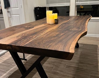 Puede incluir: Una mesa de comedor de madera con borde natural, veteado marrón oscuro y bordes irregulares. El tablero se apoya sobre una base de metal negro con un diseño angular entrecruzado. Tres velas amarillas cilíndricas están sobre la mesa.