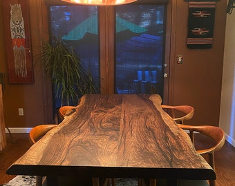 Puede incluir: Una mesa de comedor de madera con borde natural, un patrón de veta oscura y ondulada y patas de metal negro. La mesa está rodeada por cuatro sillas de madera con respaldos curvos y asientos negros, bajo una gran lámpara colgante cilíndrica.