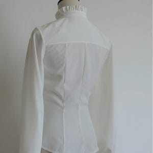 May include: White long-sleeve blouse with a ruffled collar and a fitted bodice.