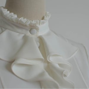 May include: A white blouse with a large, ruffled collar. The collar has multiple layers of ruffles and a single button closure.