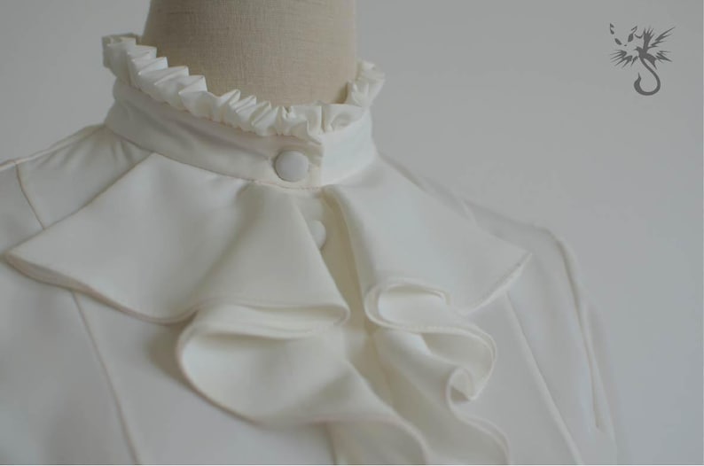 May include: A white blouse with a large, ruffled collar. The collar has multiple layers of ruffles and a single button closure.