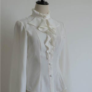 May include: A white long-sleeved blouse with a high ruffled collar and button closure. The blouse has a fitted bodice and a slightly flared hem.