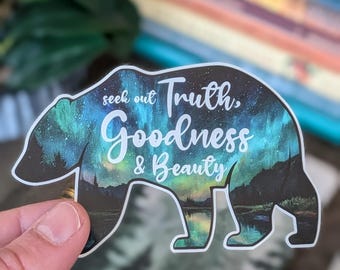 May include: A bear-shaped sticker with a landscape design featuring a lake and mountains under a starry sky. The sticker has the words "seek out Truth, Goodness & Beauty" in white and teal lettering.