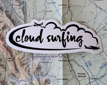 May include: A white sticker with the words "cloud surfing" in a black, handwritten font. The sticker has a black border and features airplane and helicopter silhouettes. The sticker is placed on a map.