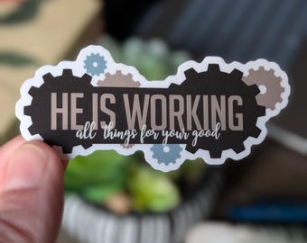 May include: A sticker featuring the words "HE IS WORKING all things for your good". The text is in white, with a black gear-shaped background and tan and blue-grey gear accents. The sticker is held by a hand.