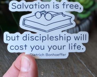 May include: A white sticker with the text "Salvation is free, but discipleship will cost you your life." The sticker features a line drawing of a book and glasses. The quote is attributed to Dietrich Bonhoeffer.