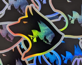 May include: A collection of holographic stickers featuring a wolf's head silhouette. The wolf's head is black with a colourful mountain and starry night scene inside. The stickers have a shiny, iridescent border.
