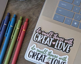 May include: A laptop with two stickers that read "do small things with GREAT LOVE." Four pens in blue, teal, green, and red are arranged next to the laptop. A decorative pot with a black and white striped design is in the background.