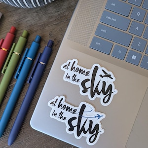 May include: Two white stickers with the text "at home in the sky" and an airplane graphic are on a laptop. Several pens in red, green, blue, and purple are arranged on a wooden surface. A small succulent plant is in a decorative pot.