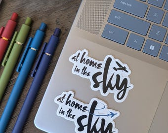 May include: Two white stickers with the text "at home in the sky" and an airplane graphic are on a laptop. Several pens in red, green, blue, and purple are arranged on a wooden surface. A small succulent plant is in a decorative pot.