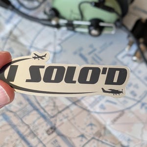 Puede incluir: Una pegatina plateada con el texto "I SOLO'D" en negro, con pequeñas siluetas de aviones. La pegatina se sostiene frente a un mapa y unos auriculares de aviación verdes.