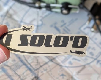 May include: A silver sticker with the text "I SOLO'D" in black, featuring small airplane silhouettes. The sticker is held in front of a map and a green aviation headset.