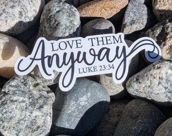 May include: A white sticker with the words "LOVE THEM Anyway" and "LUKE 23:34" in black script. The sticker is placed on a bed of smooth, grey and brown river rocks.
