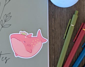 May include: A pink cartoon whale sticker with a crown on a grey notebook. A white flower and cactus in a light blue pot. Three pens in green, red, and blue on a wooden surface.