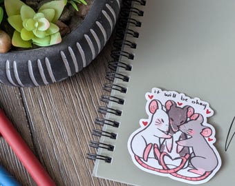 May include: A sticker showing three cartoon mice embracing, with hearts and the text "it will be okay." The sticker is on a spiral notebook next to pens and a potted succulent.