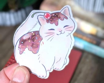 May include: A white cat sticker with a dessert theme. The cat has a chocolate frosting design with sprinkles and a cherry on top. The cat is smiling with its eyes closed. The sticker is held by a person's hand.