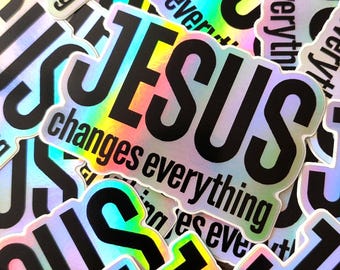 May include: A pile of stickers featuring the word "JESUS" in black, with the text "changes everything" beneath. The stickers have a shimmering, iridescent finish, displaying a spectrum of colours.