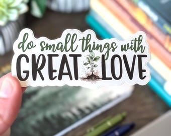 May include: A white sticker with the text "do small things with GREAT LOVE" in green and black fonts. A small plant illustration is between the words "GREAT" and "LOVE". The sticker is held by a hand, with pens and a laptop in the background.