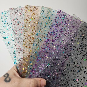 Puede incluir: Un conjunto de seis l&aacute;minas de pl&aacute;stico transparente con purpurina de diferentes colores incrustada. Las l&aacute;minas est&aacute;n dispuestas en forma de abanico y est&aacute;n siendo sostenidas por una mano.