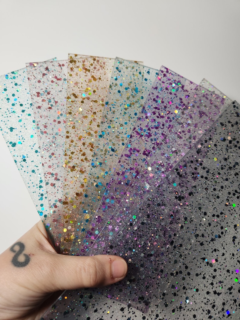 Puede incluir: Un conjunto de seis l&aacute;minas de pl&aacute;stico transparente con purpurina de diferentes colores incrustada. Las l&aacute;minas est&aacute;n dispuestas en forma de abanico y est&aacute;n siendo sostenidas por una mano.