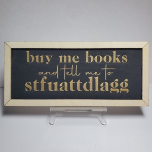 May include: A white framed sign with a black background and gold lettering. The sign reads "buy me books and tell me to stfuattddlagg".