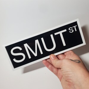 Puede incluir: Un letrero blanco y negro con el texto "SMUT ST" en letras blancas sobre fondo negro. El letrero est&aacute; enmarcado en blanco.