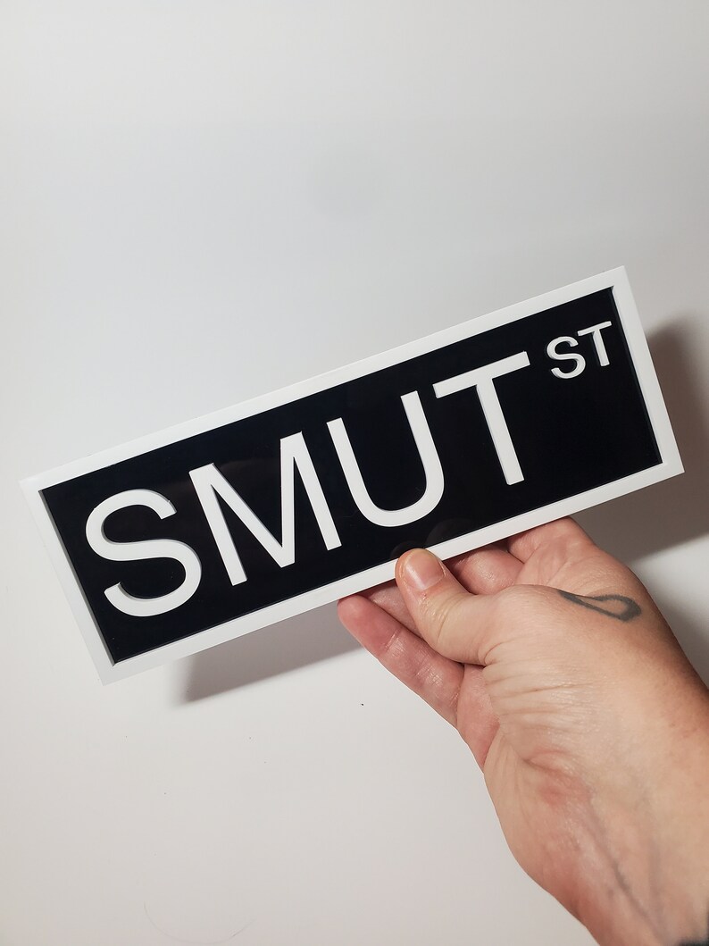 Puede incluir: Un letrero blanco y negro con el texto "SMUT ST" en letras blancas sobre fondo negro. El letrero est&aacute; enmarcado en blanco.
