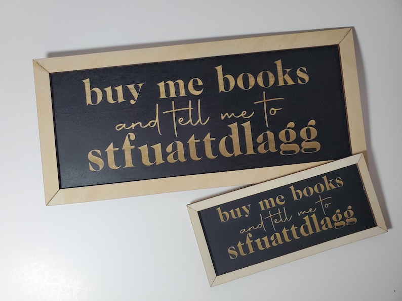 May include: Two wooden signs with black backgrounds and gold lettering. The signs read "buy me books and tell me to stfuattdlagg".