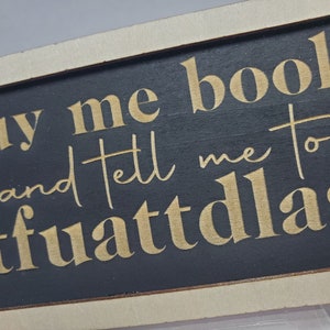 May include: A black wooden sign with gold lettering that reads "buy me books and tell me to stfuattlag".