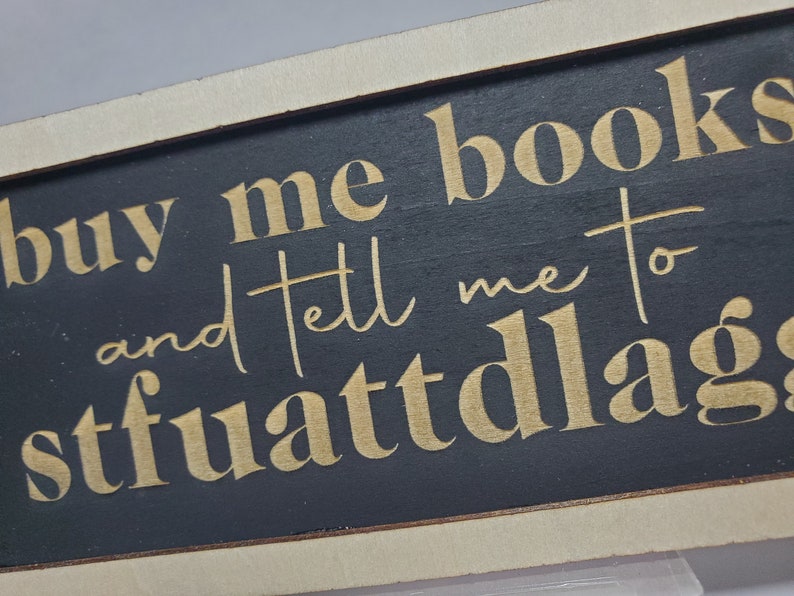 May include: A black wooden sign with gold lettering that reads "buy me books and tell me to stfuattlag".