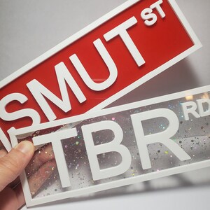 Puede incluir: Dos letreros con marco blanco y letras blancas. El letrero superior es rojo y dice "SMUT ST". El letrero inferior es transparente con un fondo brillante y dice "TBR RD".