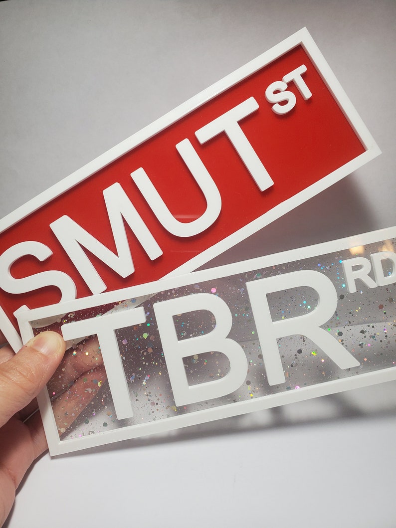 Puede incluir: Dos letreros con marco blanco y letras blancas. El letrero superior es rojo y dice "SMUT ST". El letrero inferior es transparente con un fondo brillante y dice "TBR RD".