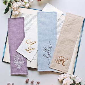 Könnte beinhalten: Vier Stoff-Lesezeichen mit gestickten Buchstaben. Die Lesezeichen sind in den Farben Lila, Blau, Beige und Weiß gehalten. Die Buchstaben beinhalten die Wörter "Bella", "A & S" und "N".