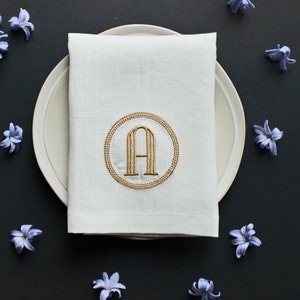 May include: A white linen napkin with a gold embroidered circle containing the letter 'A' on a white plate. The plate is surrounded by purple flowers on a black background.