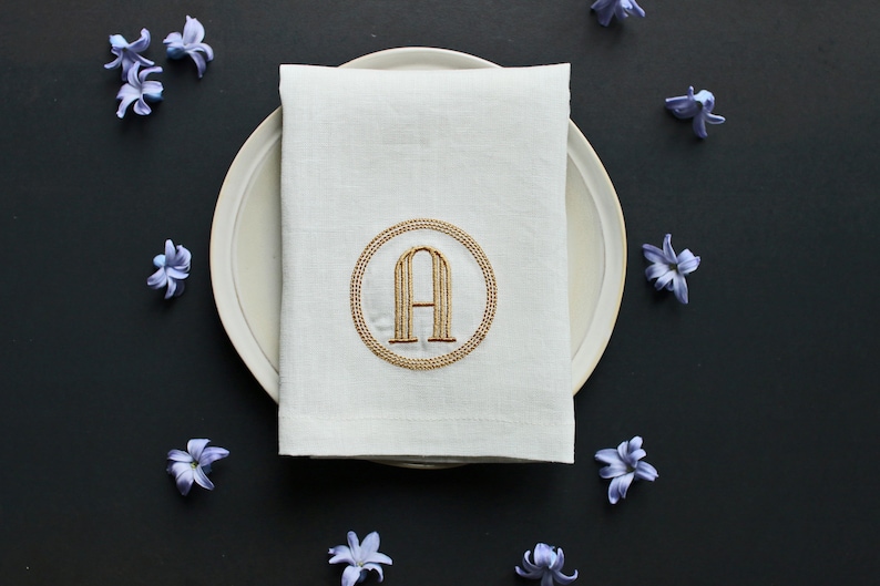 May include: A white linen napkin with a gold embroidered circle containing the letter 'A' on a white plate. The plate is surrounded by purple flowers on a black background.