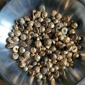 Peut inclure: Une collection de boutons en laiton antiques de différentes tailles et motifs, avec des motifs en relief et des détails décoratifs. Les boutons sont dans un bol métallique brillant, créant une esthétique vintage.