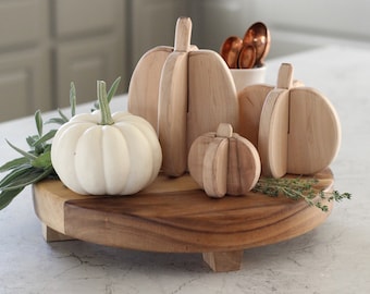 May include: A round wooden riser with three feet holds a white pumpkin, three light-colored wooden pumpkin cutouts, a small white container with copper spoons, and sprigs of green herbs. The arrangement features natural wood tones and textures.