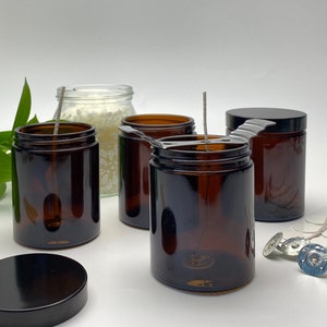 May include: A collection of amber glass jars with black lids and wicks, possibly for candle making. One jar has its lid removed, showing the inside. Additional wicks and wick centering devices are visible, indicating a DIY candle-making project.