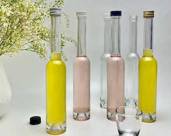 May include: Five glass bottles with different colored liquids. The bottles have silver, white, and gold caps. The bottles are on a white surface. A clear glass shot glass is in the foreground.