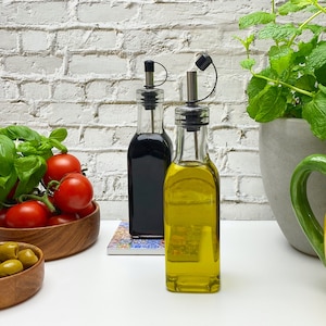 Op de afbeelding: Twee glazen flessen met zwarte en zilveren giettuiten, gevuld met olijfolie en balsamicoazijn, op een wit oppervlak met tomaten en basilicum op de achtergrond.
