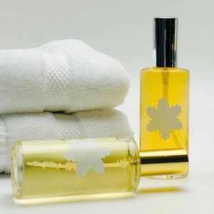 May include: Two glass bottles of body spray with a gold cap and a white snowflake design. The bottles are sitting on a white towel.