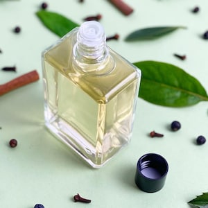 May include: A clear, square glass bottle containing a pale yellow liquid, with the cap removed. The bottle is set amongst green leaves, cloves, cinnamon sticks, and dark berries, on a pale green surface.