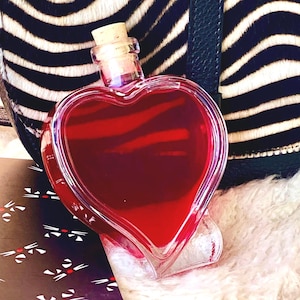 May include: A heart-shaped glass bottle filled with a deep red liquid, sealed with a cork. The bottle is set against a black and white zebra-striped pattern and a fluffy white surface.