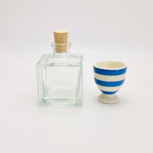 Op de afbeelding: Een heldere glazen fles met een kurken stop en een blauw-wit gestreept keramiek eierdopje.