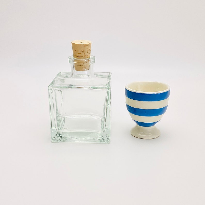 Op de afbeelding: Een heldere glazen fles met een kurken stop en een blauw-wit gestreept keramiek eierdopje.